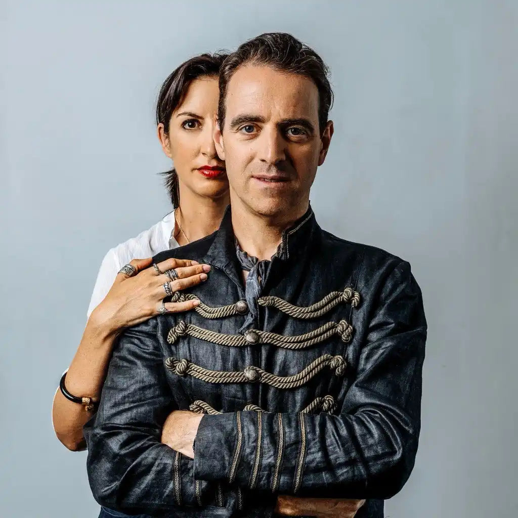 A man and a woman posing together, with the man in the foreground wearing a black, ornate military-style jacket.