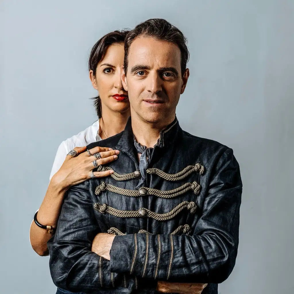 A man and a woman posing together, with the man in the foreground wearing a black, ornate military-style jacket.