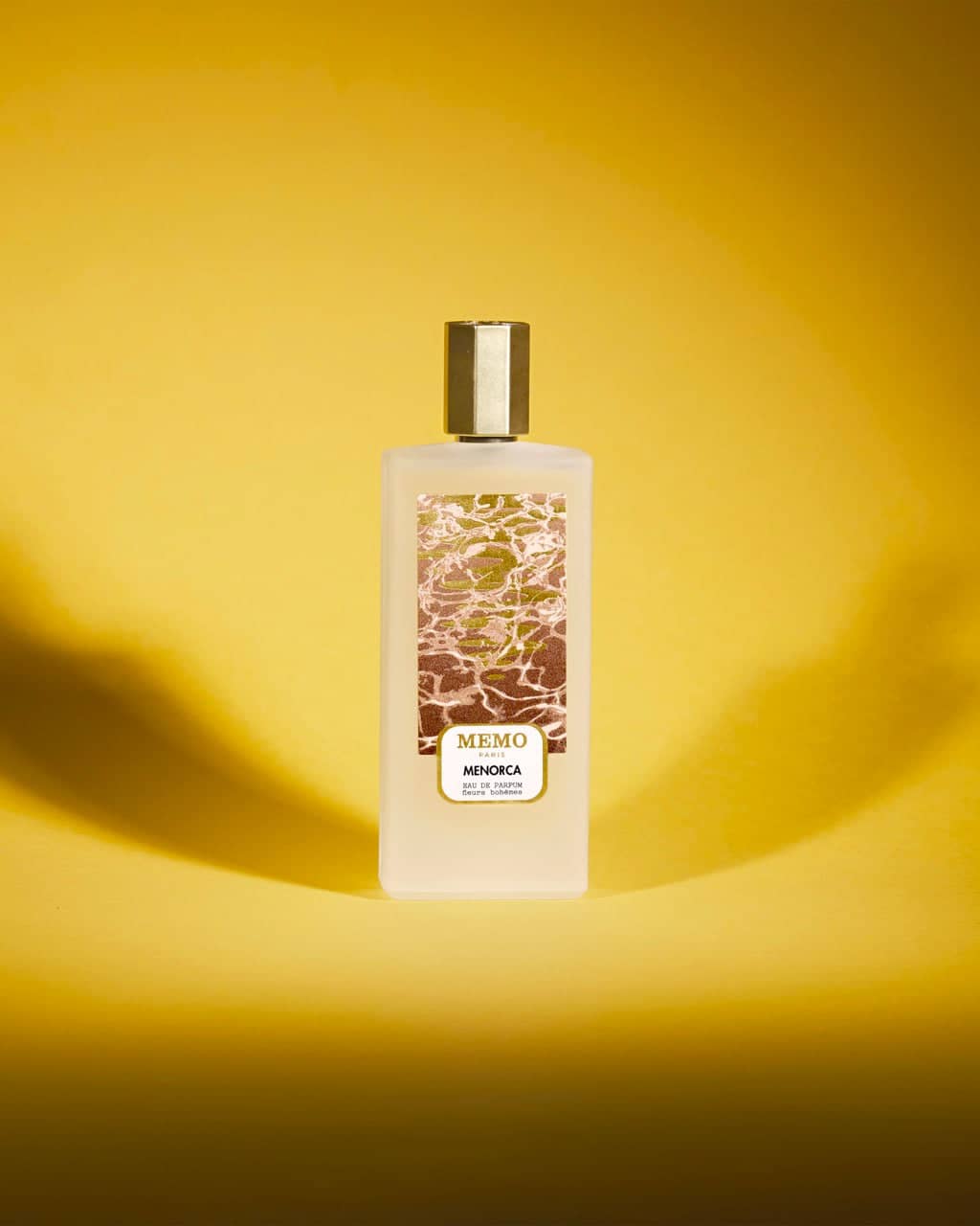 A sleek, translucent white bottle of memo menorca eau de parfum with a gold cap and an ornate label featuring a water ripple design.